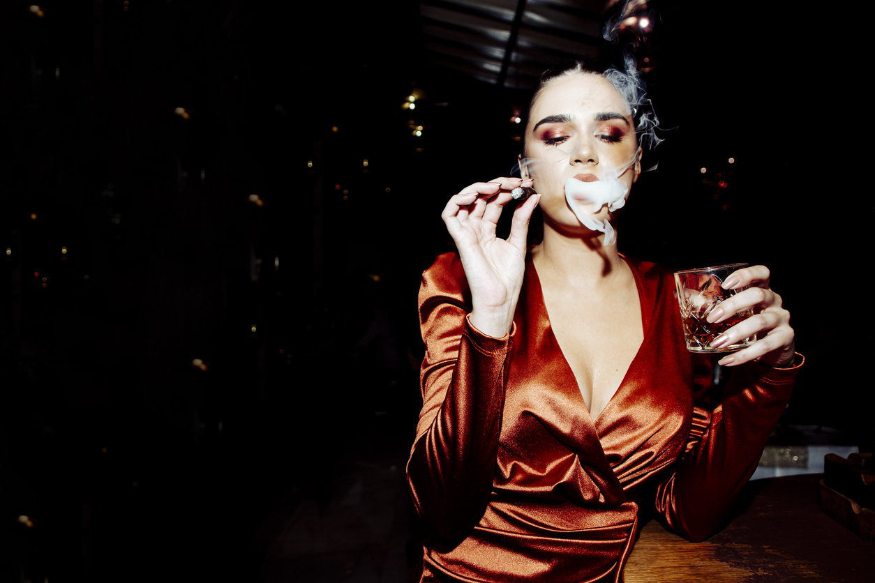 Woman in a rust satin dress exhaling cigar smoke, holding a glass of whisky