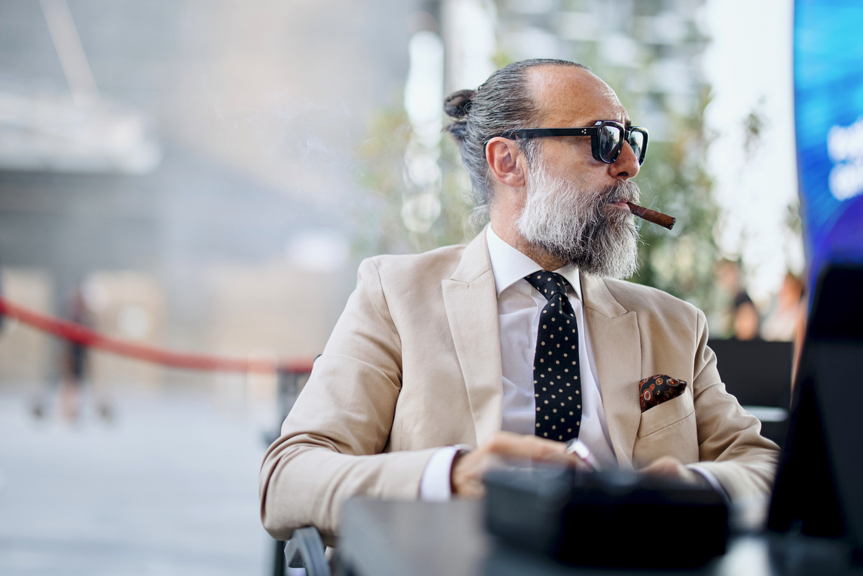 Man in a cream suit and sunglasses smoking a cigar at an outdoor table